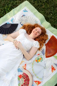Woman in a white dress lying on a patterned quilt surrounded by colorful hand fans