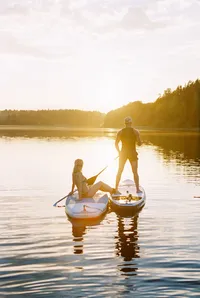 Couple paddling on a calm lake in golden light, one standing on a paddleboard and one seated in a kayak