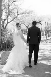 Black-and-white wedding portrait of a bride looking back while holding hands with the groom along a desert garden path.