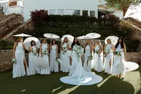 Bride and bridesmaids dressed in white posing outdoors with white parasols and bouquets.