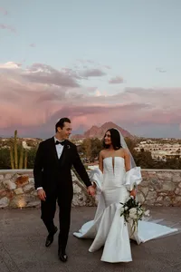 Bride and groom walking hand in hand with a desert mountain and pink evening sky behind them.