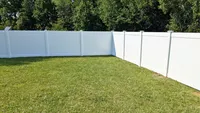 The interior corner of a bright white vinyl privacy fence surrounding a manicured green lawn, backing up to a dense tree line in the Baltimore region.