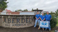 Blue Crab Fence & Home owners Jim, Brent, and Brian stand with their yard sign in front of the Aberdeen All America City welcome sign.