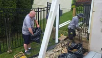Two workers properly setting a white vinyl fence post by pouring high-strength concrete mix and tamping it securely into a Maryland yard.