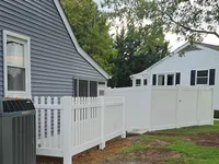 Newly installed four-foot white vinyl picket fence transitioning into a six-foot privacy fence in a Lutherville-Timonium, Maryland backyard. The low-maintenance PVC picket section cleanly borders the side of a grey residential home and an air conditioning unit.