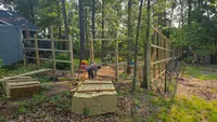 A massive stack of fresh premium lumber staged in a shaded yard while a worker builds a tall custom wood fence frame in the Baltimore area.