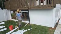 A Blue Crab Fence installer building custom white vinyl privacy panels directly underneath a raised wooden deck to enclose the lower yard space of a Maryland home.
