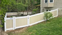 A sloped, two-tone tan and white vinyl privacy fence with a secure gate, following the natural grade of a wooded backyard in the Baltimore region.