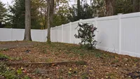 A solid white vinyl privacy fence following the natural slope of a wooded backyard in the Baltimore region, installed over a leaf-covered landscape.