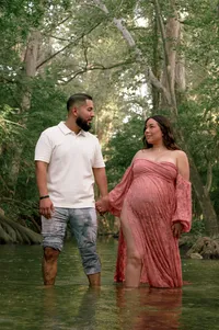 Expectant parents hold hands while standing in a creek and looking at each other.