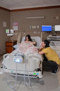 Dad kneels beside the hospital bed while Mom breastfeeds their newborn.