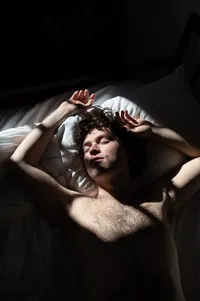 A person reclines on a bed with their arms above their head in dramatic window light.