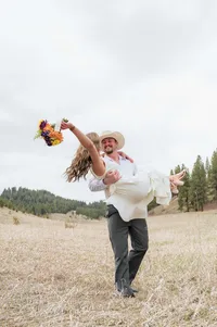 One wedding partner carries the other through a grassy field as they raise a colorful bouquet.