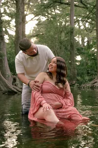 Expecting parents sitting in a creek as dad holds her shoulders.