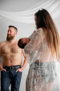Postpartum mother bottle feeds her newborn as her husband stands shirtless in the background.