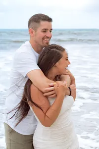 Man holding his fiancé as they laugh and stare into the sea.