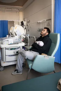 Tired dad sits in a hospital chair and holds his newborn.