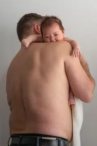 shirtless postpartum father holds his son to his chest.