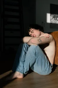 Non-Binary person resting their head on their knees as they bask in the window light.