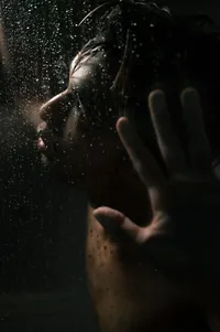 Queer man looking up toward the dim shower light as he rest his hand on the shower glass.
