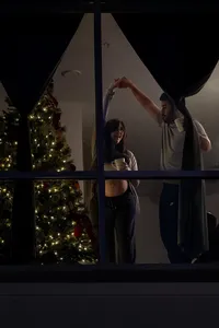 Expectant parents dance indoors, photographed through the window from outside.