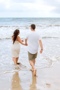 An engaged couple running into the ocean, hand in hand.