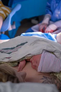 New mother holds her freshly birthed baby to her chest while doctors clean up in the background.