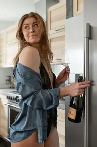 A person in an open blue shirt holds a stemmed glass beside a refrigerator.
