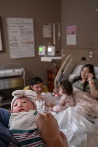 Newborn baby looks at the person holding her while her parents and sister rest on the hospital bed in the background.