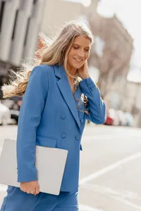 Lucy Saarelaht in a blue suit walks outdoors, holding a white folder and touching her hair; warm sunlight gives the city scene a confident, upbeat feel.