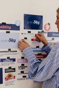 A woman in a striped shirt arranges magazine and newspaper clippings on a wall, creating a colorful media collage in a focused, creative mood.