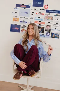 Smiling woman sits curled in a white chair, wearing a blue striped shirt and burgundy pants beneath a wall of magazine covers; relaxed, confident mood.