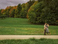 An engaged couple embraces in a wide green meadow bordered by trees.