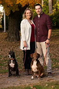 An engaged couple stands with their two dogs beneath autumn leaves.
