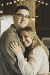 Mike and Molly smiling in a close embrace under string lights in a rustic barn during their engagement session