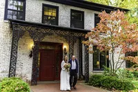 Bride and groom standing in front of a stone house with a red door beside an autumn maple tree