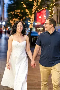 Engaged couple walking hand in hand down a brick city sidewalk lined with trees wrapped in twinkle lights