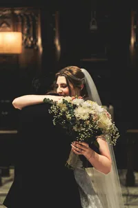 Bride in her veil hugging a loved one tightly inside a softly lit church, her bouquet in hand