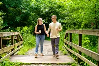 Rachel and John walking hand in hand across a wooden footbridge surrounded by green trees