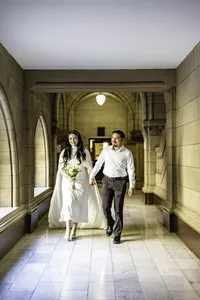 Bride holding yellow flowers and groom walking hand in hand down a long arched stone hallway