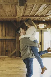 Mike lifting Molly as she wraps her arms around him and kicks up one foot in a sunlit barn