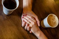 Engaged couple holding hands on a wooden café table between two coffee cups, her engagement ring in focus
