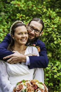 Groom hugging his smiling bride from behind as she holds a bouquet of peach and coral roses