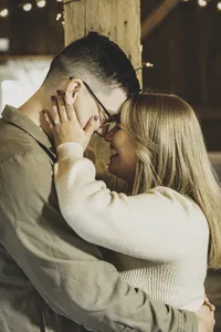 Mike holding Molly's face as they lean in close beneath strands of string lights in a wooden barn