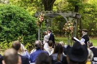 Bride and groom facing each other under a flower-covered garden arbor as guests watch their outdoor ceremony