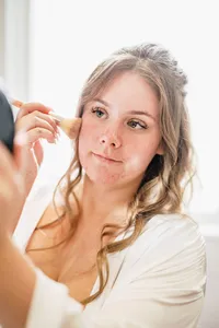 Bride in a white robe looking up as her makeup is brushed on during the wedding morning