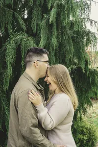 Mike kissing Molly's forehead as she laughs with her hand on his shoulder beneath a tall evergreen tree