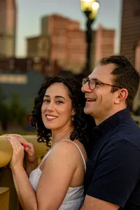 Couple smiling together against a yellow bridge railing with city buildings softly blurred behind them