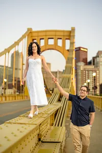 Rebecca in a white dress on the railing of the Roberto Clemente Bridge as Jordan holds her hand
