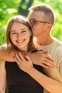 Smiling woman showing her engagement ring as her fiancé hugs her from behind among green trees